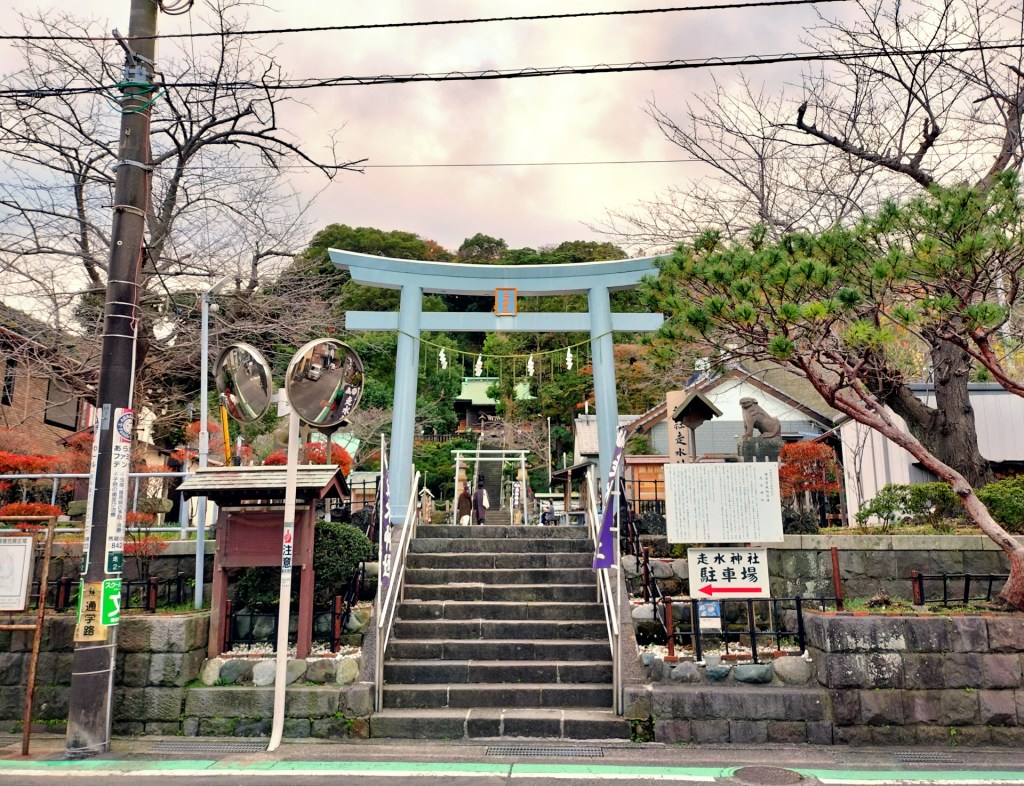走水神社の鳥居と階段の風景。神社の入口には狐の像があり、周囲には木々や花が点在している。