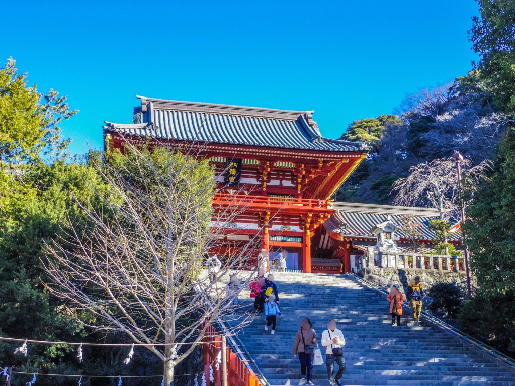 鶴岡八幡宮の赤い門と階段を上る人々の風景。青空の下で緑に囲まれた神社の景観。